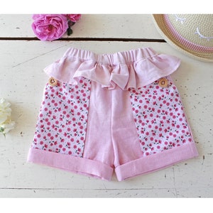 Peut inclure: Short en tissu rose avec une ceinture à volants et des panneaux en tissu à imprimé floral. Le short a des boutons en bois et est fait d'un tissu doux et léger.