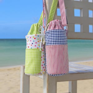 Pode incluir: Dois porta-garrafas de tecido com fechos de cordão pendurados numa cadeira branca. Um é xadrez verde e bolinhas, o outro é xadrez azul e listras vermelhas. O fundo é uma cena de praia.