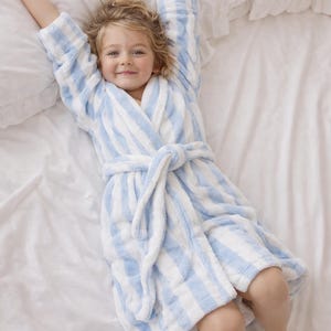May include: A child's blue and white striped bathrobe. The soft, plush robe is tied at the waist and the child is lying on a white bed. The child is smiling and has their arms stretched above their head.