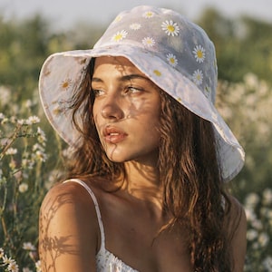 May include: A white bucket hat featuring a sheer overlay with an embroidered daisy pattern. The hat has a wide, flexible brim and a delicate floral design with white petals and yellow centers scattered across the crown and the underside of the brim.