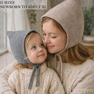 May include: Two knit bonnets with pointed tops and chin ties. One bonnet is heather gray, and the other is a light beige. The text 11 SIZES NEWBORN TO ADULT XL is visible in the upper left corner. These head coverings feature a textured knit fabric.