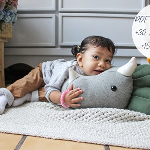 May include: A young child is lying on a light gray rug, hugging a gray and green plush dinosaur toy. The toy has a white horn and black eyes. The child is wearing a gray sweater, brown pants, and white socks. The text "PDF PATTERN +30 PICTURES +15 PAGES" is visible in the upper right corner of the image.