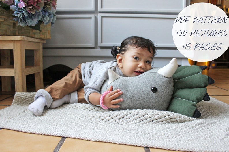 May include: A young child is lying on a light gray rug, hugging a gray and green plush dinosaur toy. The toy has a white horn and black eyes. The child is wearing a gray sweater, brown pants, and white socks. The text "PDF PATTERN +30 PICTURES +15 PAGES" is visible in the upper right corner of the image.