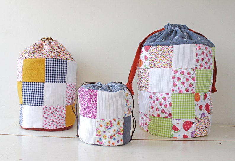 May include: Three fabric drawstring bags in different sizes. The largest bag is blue and white with a patchwork design. The medium bag is pink, white, and blue with a patchwork design. The smallest bag is yellow, white, and blue with a patchwork design.