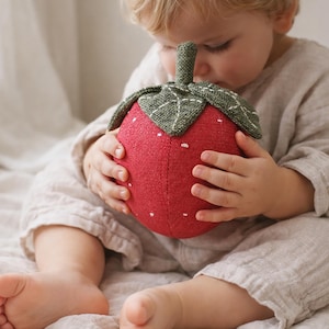 Puede incluir: Un juguete de fresa de peluche rojo con detalles de semillas blancas y hojas verdes. El juguete lo sostiene un niño que lleva un atuendo de lino de color claro. El juguete de fresa es un artículo suave y táctil.