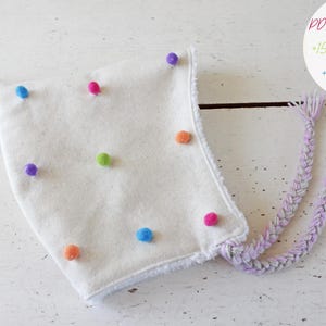 Op de afbeelding: Een witte muts met kleurrijke pompons en gevlochten banden. De muts is versierd met verschillende gekleurde pompons, waaronder groen, blauw, roze, oranje en paars. De tekst op de afbeelding luidt "PDF PATTERN +15 PICTURES +8 PAGES".