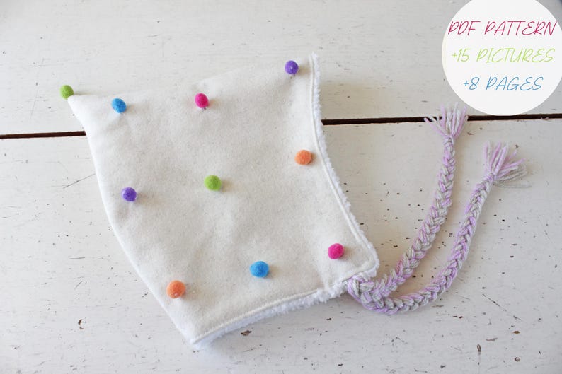 Op de afbeelding: Een witte muts met kleurrijke pompons en gevlochten banden. De muts is versierd met verschillende gekleurde pompons, waaronder groen, blauw, roze, oranje en paars. De tekst op de afbeelding luidt "PDF PATTERN +15 PICTURES +8 PAGES".