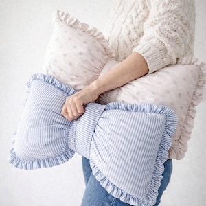 Könnte beinhalten: Ein dekoratives kissenförmiges Kissen mit blau-weißen Streifen wird im Bild gehalten. Das Kissen hat einen gerüschten Rand. Ein weiteres Kissen mit rosa Hintergrund und kleinen weißen Punkten ist ebenfalls sichtbar.