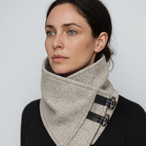 May include: A beige herringbone patterned scarf with black leather straps and silver-tone buckles. The scarf is wrapped around the neck and fastened with two buckles. The person is wearing a black top.