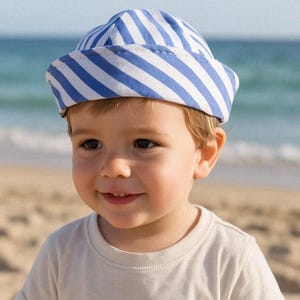 Può includere: Un cappello da marinaio a righe blu e bianche. Il cappello è indossato da un bambino piccolo. Il bambino indossa una maglietta bianca e sorride. Lo sfondo è una spiaggia con l'oceano in lontananza.