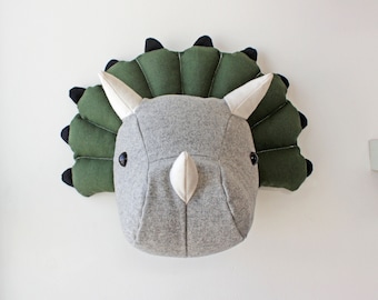 May include: A gray and green plush triceratops head wall decor. The head is made of soft fabric and has a textured green frill around the neck. The eyes are black plastic buttons.