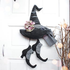 May include: A black witch's hat with a gray ribbon and a pink flower, hanging on a white door with a pair of black witch's shoes hanging below.
