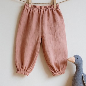 Op de afbeelding: Zachtroze babybroek met een elastische tailleband en gerimpelde enkels, hangend aan een waslijn met houten knijpers. Een grijs stoffen eendenspeelgoed staat op een houten oppervlak onder de broek.