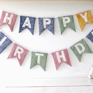 May include: A colorful "Happy Birthday" banner with fabric pennants in pink, blue, green, and yellow. Each pennant displays a white letter. A stuffed bunny sits below the banner, creating a festive scene.