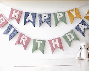 May include: A colorful "Happy Birthday" banner with fabric pennants in pink, blue, green, and yellow. Each pennant displays a white letter. A stuffed bunny sits below the banner, creating a festive scene.
