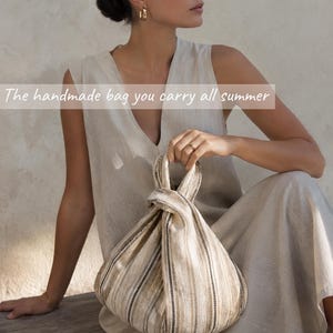 May include: A beige and brown striped fabric bag with a knotted handle design. The bag features vertical stripes in varying shades of tan, cream, and dark brown. The text "The handmade bag you carry all summer" is overlaid across the center of the image.