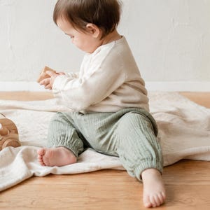Pode incluir: Uma criança pequena vestindo uma camisa de manga comprida cor de creme e calças de musselina verde sálvia, segurando um pequeno bloco de madeira. Ao lado da criança, um veículo de brinquedo de madeira natural com recortes sobre um tecido claro.