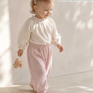 May include: A long-sleeved cream-colored blouse with a ruffled collar and elastic cuffs paired with light pink elastic-waist trousers. The text "MADE FOR LITTLE ADVENTURES" is visible in the upper right corner. A small wooden toy is held in one hand.