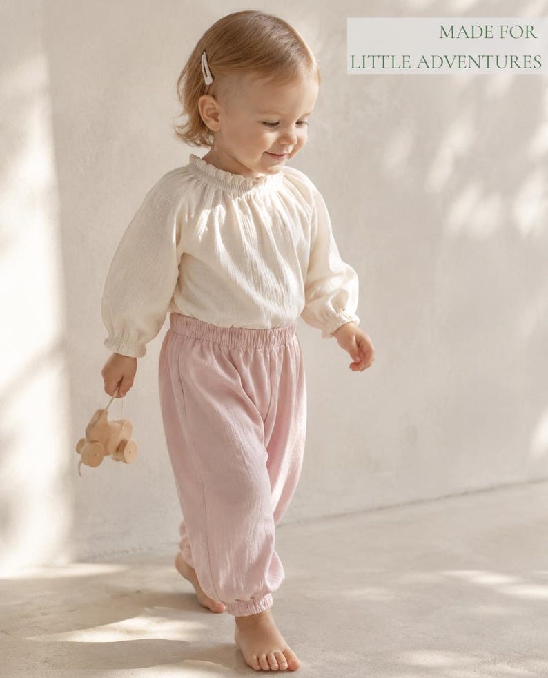 May include: A long-sleeved cream-colored blouse with a ruffled collar and elastic cuffs paired with light pink elastic-waist trousers. The text "MADE FOR LITTLE ADVENTURES" is visible in the upper right corner. A small wooden toy is held in one hand.