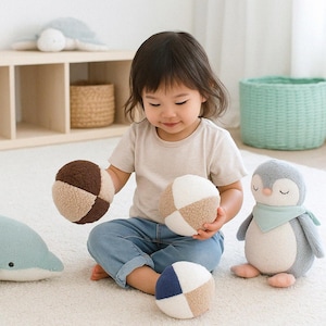 May include: A child plays with soft toys, including three colour-blocked balls, a dolphin, and a penguin. The balls have brown, beige, and blue sections. The penguin has a blue scarf. The toys are on a white rug.