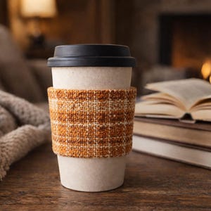 Könnte beinhalten: Ein beiger Reisebecher mit schwarzem Deckel und einer gewebten orange-braunen Manschette. Die Tasse steht auf einer Holzoberfläche, im Hintergrund Bücher und eine Decke. Ein Kamin ist zu sehen.