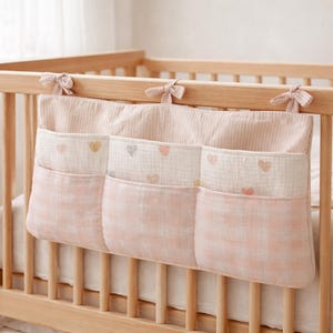 以下が含まれることがあります： 木製のベビーベッドに取り付けられた、ピンクと白の保育園オーガナイザー。3つのポケットがあり、ポケットにはギンガムチェック柄とハート型のアクセントが付いています。オーガナイザーは小さなリボンで固定されています。