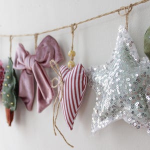 Può includere: Una ghirlanda di ornamenti in tessuto di varie forme e colori. Include un cuore rosa, un albero verde, un fiocco malva, un cuore a righe, una stella con paillettes e un cuore verde, sospesi a uno spago.
