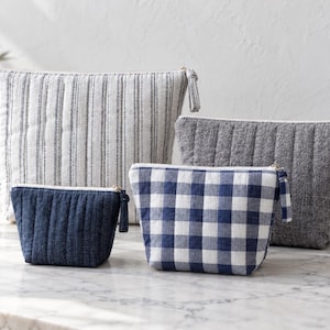 Peut inclure: Un ensemble de quatre pochettes matelassées de tailles variées. Une grande pochette présente un motif à rayures verticales blanches et grises, une moyenne est gris uni, une autre à carreaux vichy bleu et blanc, et une petite est bleu marine uni.