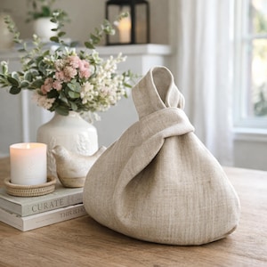 May include: A beige linen knot bag with a single handle formed by tying two fabric ends together. The bag features a textured, woven appearance in a neutral light tan color, showcasing a simple, unstructured shape that rests on a flat surface.
