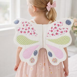 May include: White butterfly wings with strawberry and gingham patterns. The wings have pink, green, and blue accents. The wings are worn by a child wearing a pink dress.