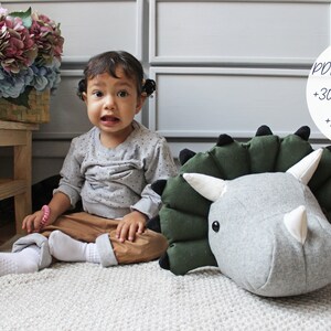 May include: A child sits on a white rug next to a large gray and green plush dinosaur head. The dinosaur has black eyes and a white horn. The text "PDF PATTERN +30 PICTURES +15 PAGES" is visible in the background.