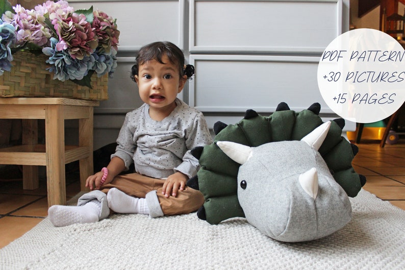 May include: A child sits on a white rug next to a large gray and green plush dinosaur head. The dinosaur has black eyes and a white horn. The text "PDF PATTERN +30 PICTURES +15 PAGES" is visible in the background.