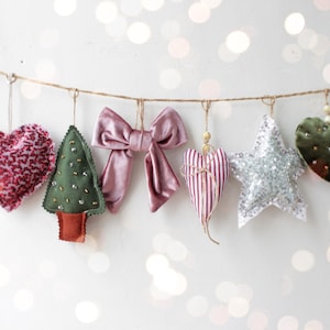 Może przedstawiać: Kolekcja ręcznie robionych ozdób świątecznych zawieszonych na sznurku. Ozdoby obejmują różowe serce, zielone drzewko, różową kokardę, serce w paski, srebrną gwiazdę z brokatem i zielone serce. Świąteczny wystrój.