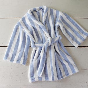 May include: A blue and white striped bathrobe with a matching belt. The robe is made of a soft, absorbent material and is laid out on a white wooden surface. The robe has a shawl collar and long sleeves.