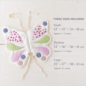 May include: Quilted butterfly wings with strawberry, gingham, and floral patterns. Text reads: THREE SIZES INCLUDED. Small: 13" x 15" (1-2 years). Medium: 14" x 16" (3-5 years). Large: 15" x 17" (6+ years). Includes cream-colored fabric ties.