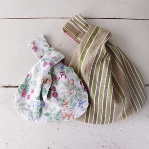 May include: Two fabric knot bags with a wrist handle design. One bag features a white background with a colorful pattern of berries, leaves, and nuts. The second bag displays a vertical striped pattern in shades of beige, olive green, and brown.