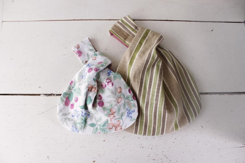 May include: Two fabric knot bags with a wrist handle design. One bag features a white background with a colorful pattern of berries, leaves, and nuts. The second bag displays a vertical striped pattern in shades of beige, olive green, and brown.