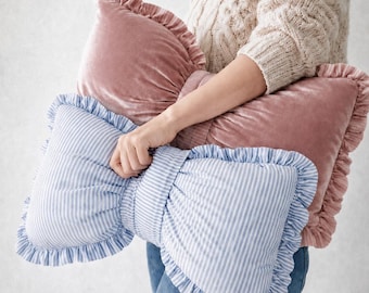 May include: A bow-shaped throw pillow featuring a dusty pink velvet section and a light blue and white striped cotton section. Both sides of the pillow are finished with matching ruffled fabric edges, creating a soft, decorative accent piece.