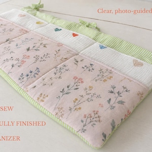 May include: A quilted crib organizer featuring a light pink fabric with a delicate floral print, a white textured section with small embroidered hearts, and green striped trim with fabric ties. Text reads: LEARN TO SEW A BEAUTIFULLY FINISHED CRIB ORGANIZER and Clear, photo-guided instructions.