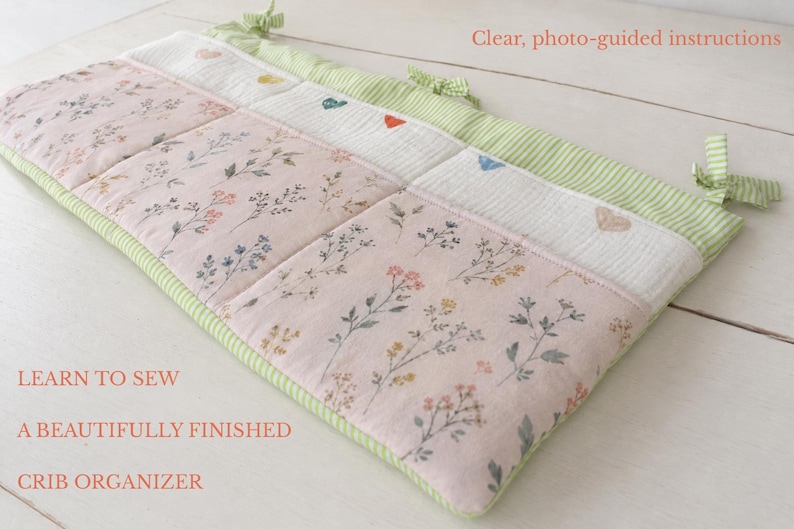 May include: A quilted crib organizer featuring a light pink fabric with a delicate floral print, a white textured section with small embroidered hearts, and green striped trim with fabric ties. Text reads: LEARN TO SEW A BEAUTIFULLY FINISHED CRIB ORGANIZER and Clear, photo-guided instructions.