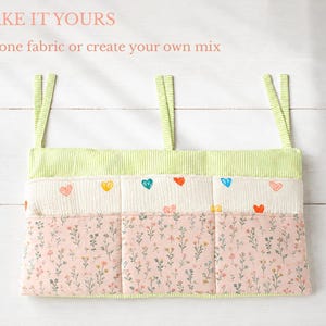 May include: A fabric wall organizer with three pockets featuring three hanging straps. The top section has lime green vertical stripes, the middle section is cream with small colorful hearts, and the bottom section is pink with a floral print. Text reads: MAKE IT YOURS. Use one fabric or create your own mix.
