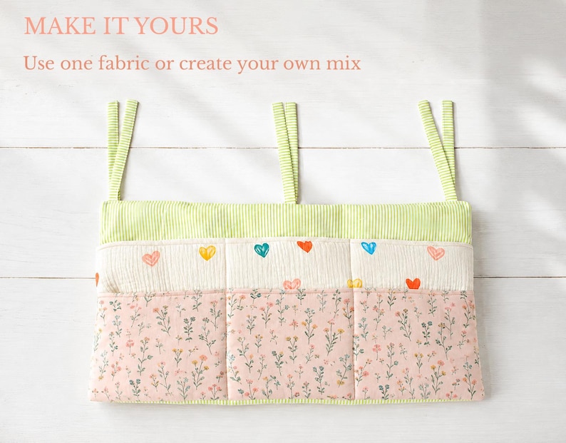 May include: A fabric wall organizer with three pockets featuring three hanging straps. The top section has lime green vertical stripes, the middle section is cream with small colorful hearts, and the bottom section is pink with a floral print. Text reads: MAKE IT YOURS. Use one fabric or create your own mix.