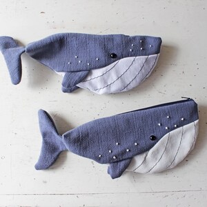 Pu&ograve; includere: Due astucci in tessuto a forma di balena di un colore grigio-blu testurizzato. Ogni astuccio presenta una parte inferiore bianca con linee verticali cucite, un occhio a perla nera e piccoli punti ricamati bianchi sulla parte superiore.