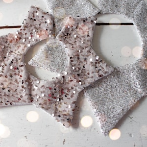 May include: Close-up of several star-shaped ornaments. The ornaments are covered in silver sequins and small, shiny embellishments. The stars are arranged on a white surface, creating a festive and decorative display.