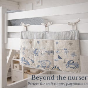 May include: A hanging bedside storage organizer featuring three pockets with a light beige fabric base, blue dinosaur illustrations, and small heart motifs. The top section has a blue and white pinstripe pattern and three fabric ties for attachment.