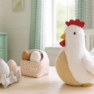 Könnte beinhalten: Ein weißes Stoffhuhn mit rotem Kamm und Kehllappen sowie gelbem Schnabel sitzt neben einem Karton mit Stoffeiern. Ein kleiner Blumenkorb enthält zusätzliche Stoffeier. Die Szene spielt auf einer hellen Holzoberfläche.