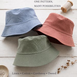May include: Three bucket hats in blue, terracotta, and sage green. The text "ONE PATTERN, MANY POSSIBILITIES" and "Linen • Cotton • Corduroy • Tweed" is visible. A wooden spool of thread is positioned near the top right corner of the hats.