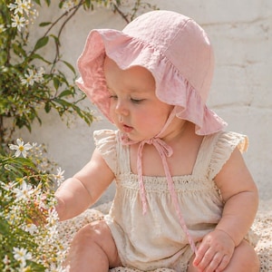 Puede incluir: Un bebé con un gorro rosa claro con un borde con volantes y una cinta a juego. El bebé también lleva un pelele color crema con detalles de encaje. El bebé está sentado en una manta color crema y alcanza flores blancas.