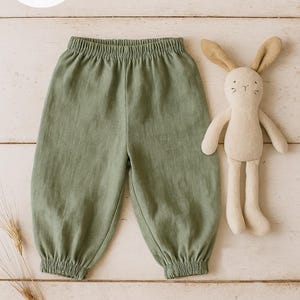 May include: Sage green pants with an elastic waistband and gathered elastic cuffs, accompanied by a beige plush rabbit toy. The image includes text that reads: PDF PATTERN +20 PICTURES +15 PAGES.