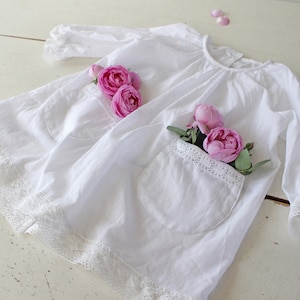 Op de afbeelding: Een witte katoenen jurk met kant en twee zakken. Roze rozen zijn in de zakken geplaatst.